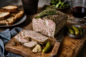 Pâté de campagne maison tranché sur une planche en bois, entouré de cornichons, de pain de campagne et d'aromates frais